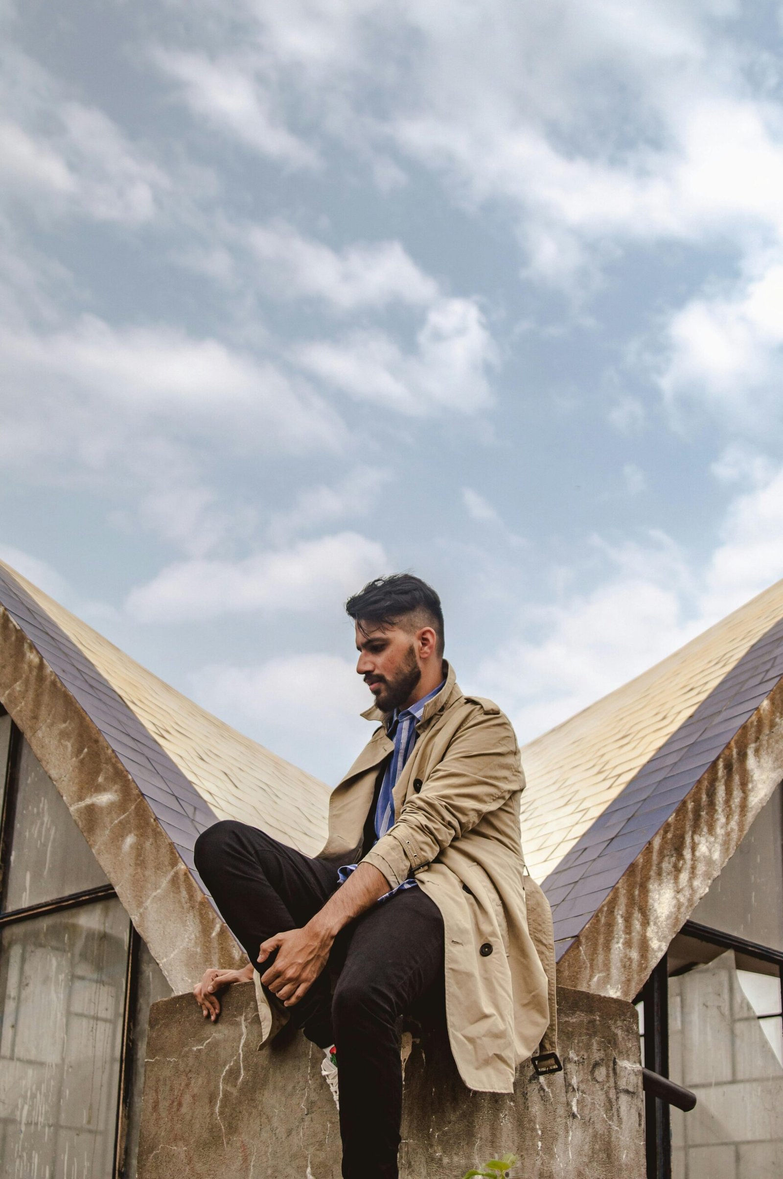 Fashionable young man with a full beard sitting on a modern urban structure outdoors.
